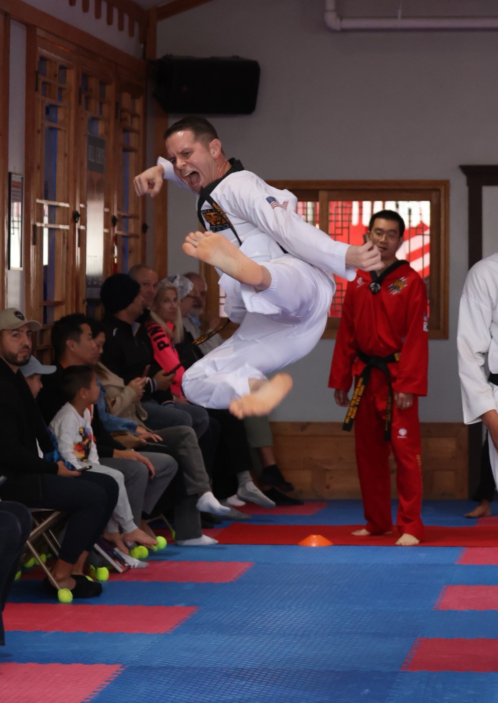 Adult student performing a flying kick during a belt test as families watch at Bill Cho's dojang