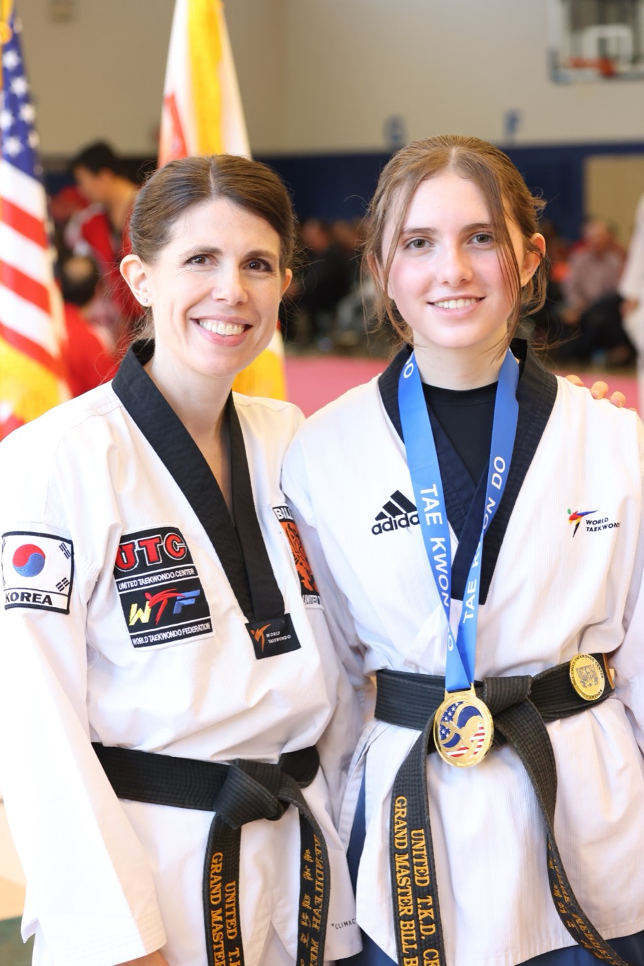 Mother and teen daughter in black belts at a tournament, the daughter wearing a gold medal